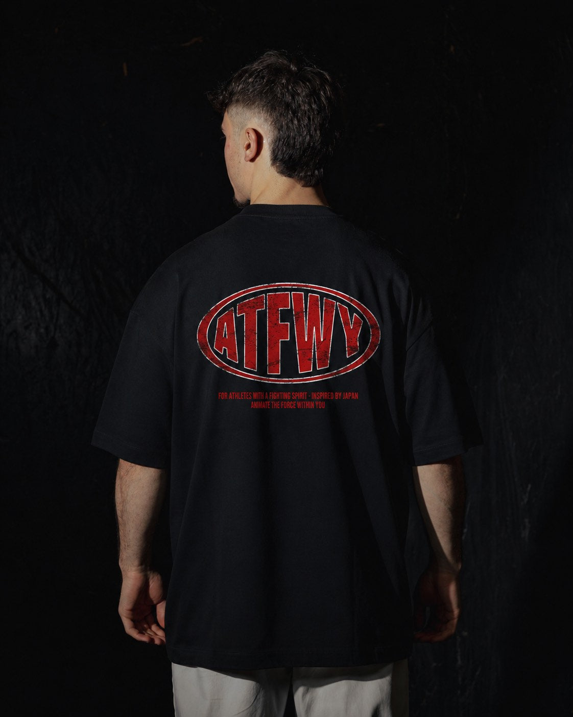 Homme de dos portant un t-shirt oversize de couleur noire, avec un visuel des lettres ATFWY en rouge, destiné aux athlètes et inspiré de la culture japonaise.