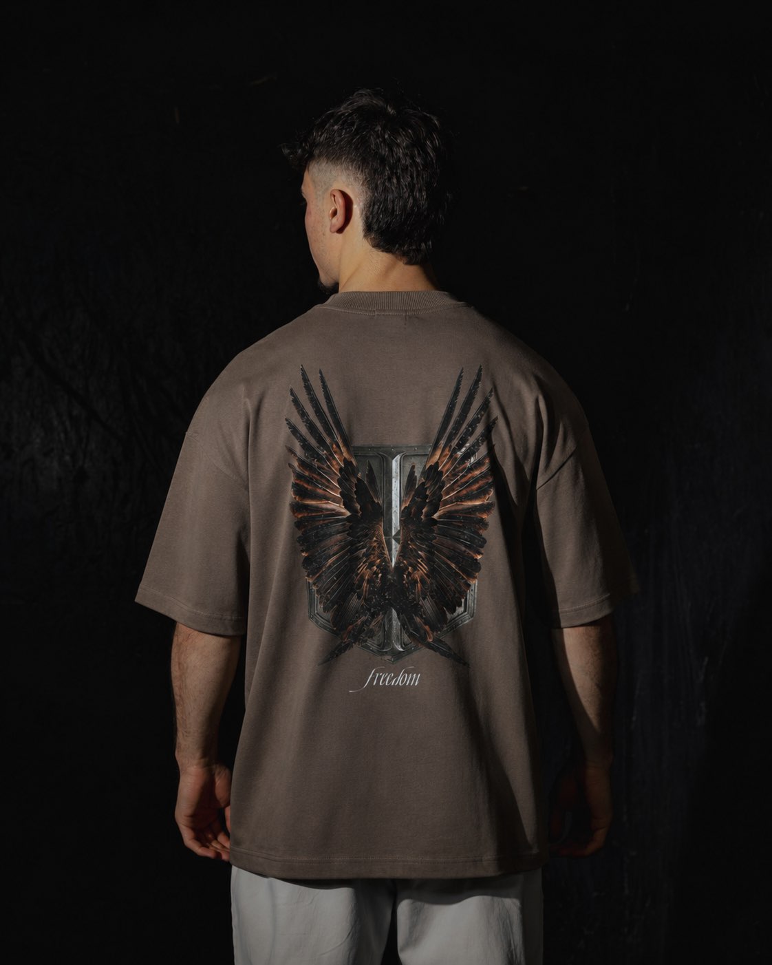 Homme de dos portant un t-shirt oversize de couleur mocha, avec un visuel inspiré de l’anime l’Attaque des Titans, représentant un bouclier et des ailes.