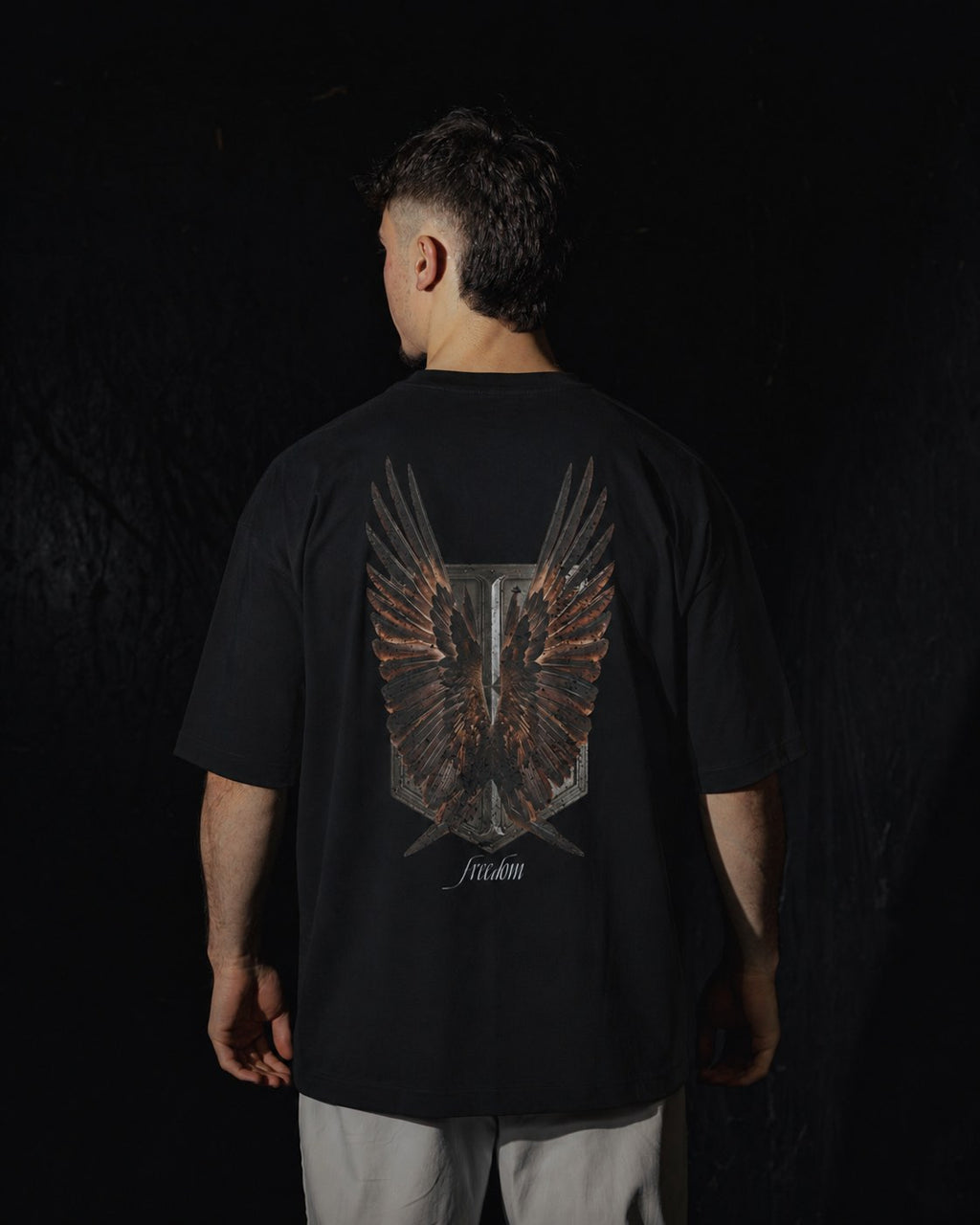 Homme de dos portant un t-shirt oversize de couleur noire, avec un visuel inspiré de l’anime l’Attaque des Titans, représentant un bouclier et des ailes.